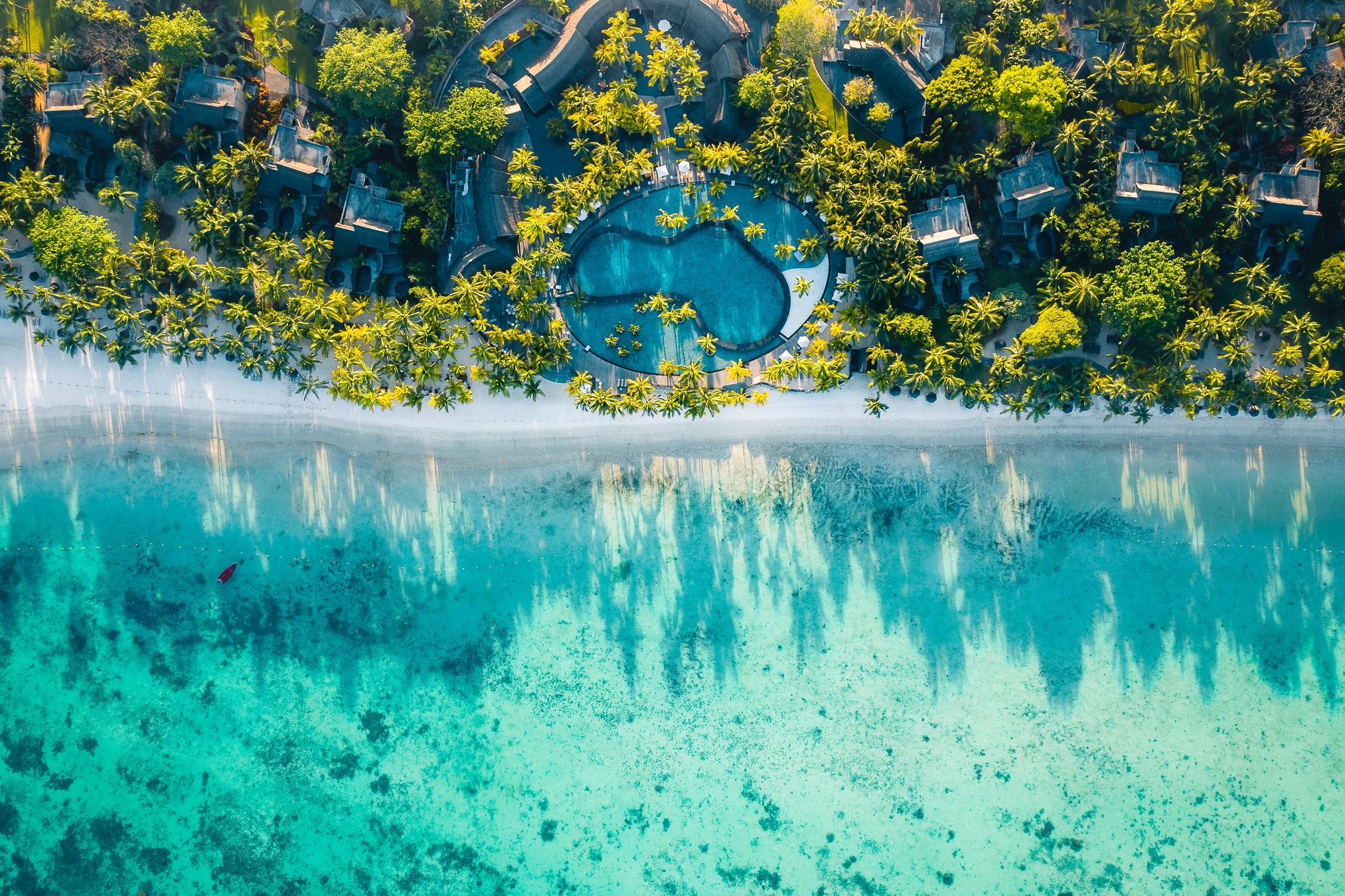 Vista aerea di un resort tropicale sulla spiaggia con acqua turchese, sabbia bianca, palme verdi e lussureggianti, ville e una piscina centrale circondata dal verde.