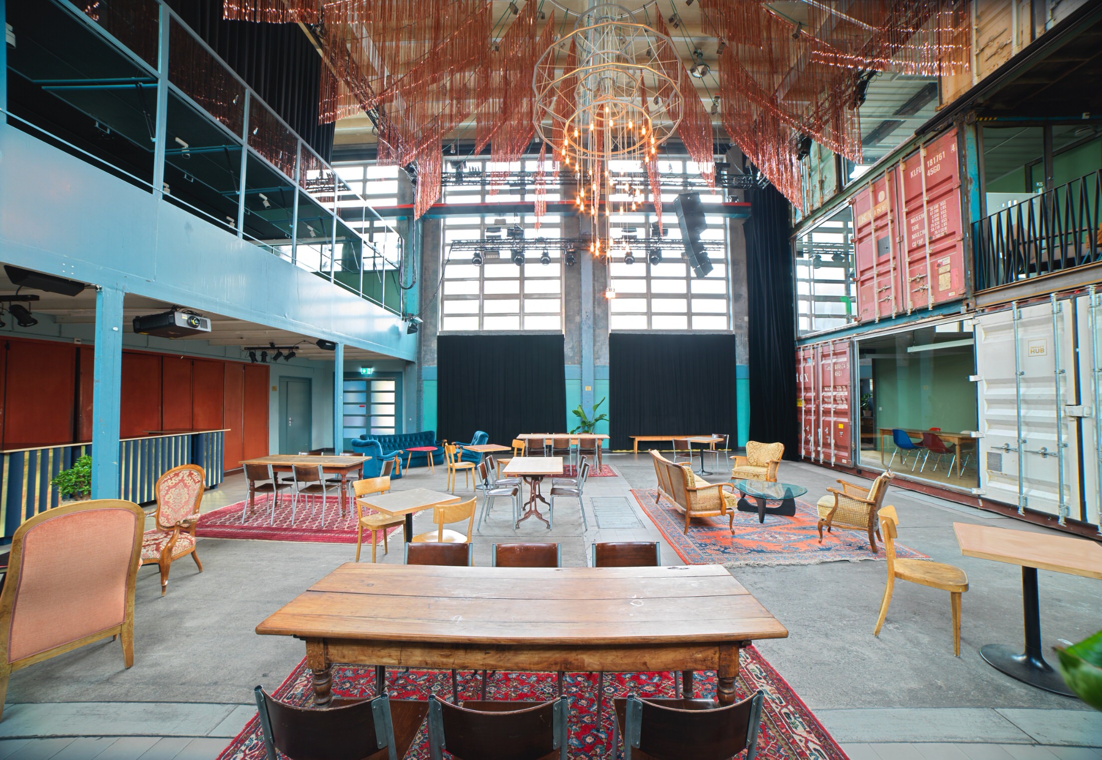 Ampio locale moderno in stile industriale a Lugano con mobili vintage eclettici, tavoli in legno, tappeti colorati, grandi finestre, travi a vista, container navali e un lampadario sospeso fatto di tubi di rame.