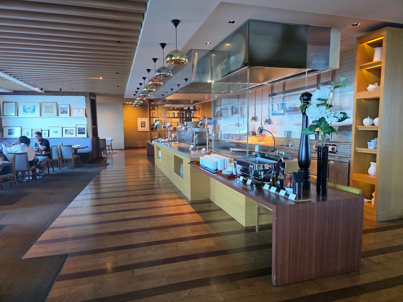 A modern buffet setup in a spacious restaurant with wooden floors, an open kitchen, food warmers, decorative shelves, and diners seated in the background near artworks on the wall.