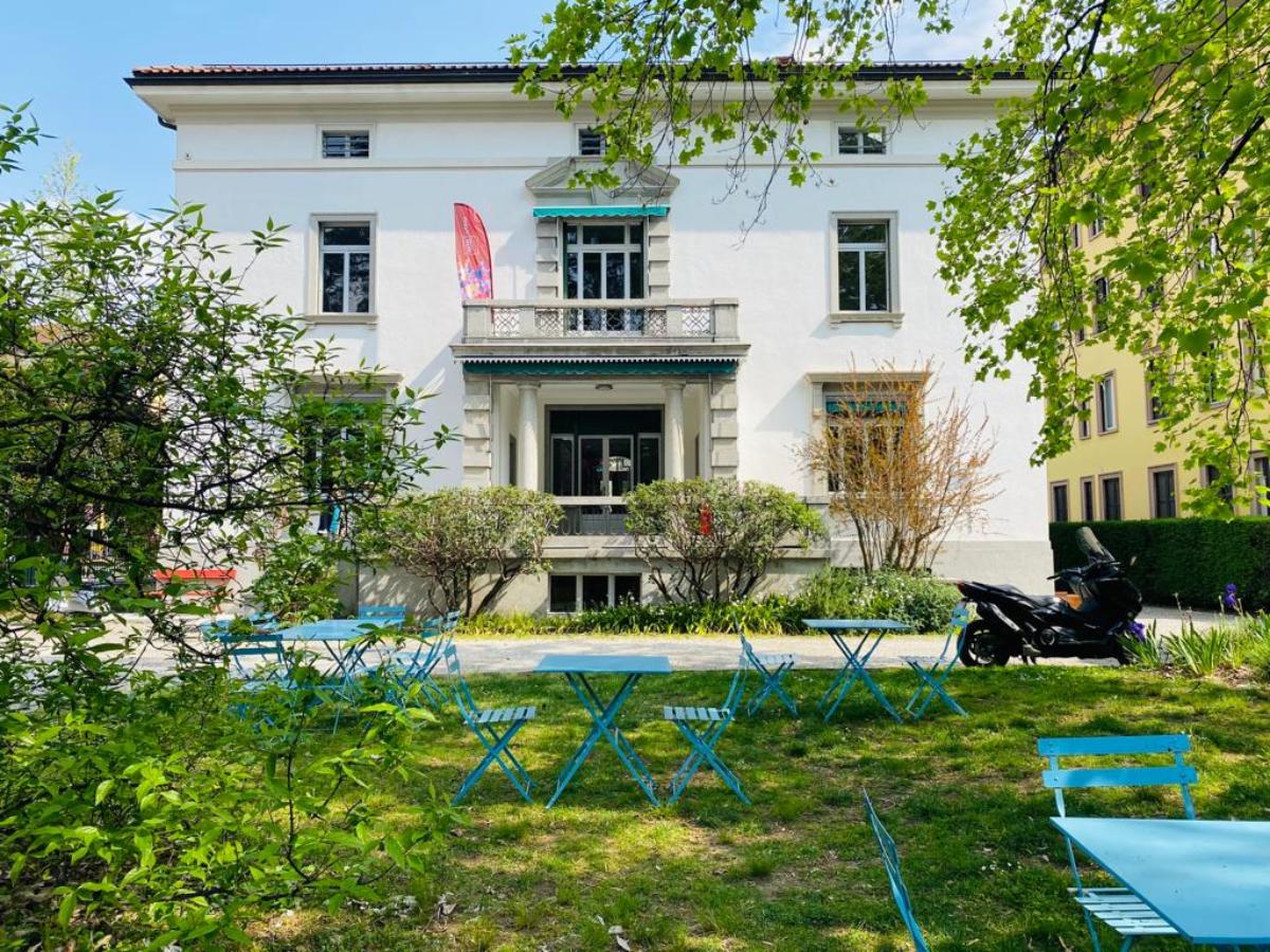 Un edificio bianco a due piani di ispirazione reggiana con un piccolo balcone e una tenda da sole blu, circondato da alberi e cespugli verdi. Sul prato erboso in primo piano sono disposti tavoli e sedie blu. Una moto è parcheggiata sulla destra.