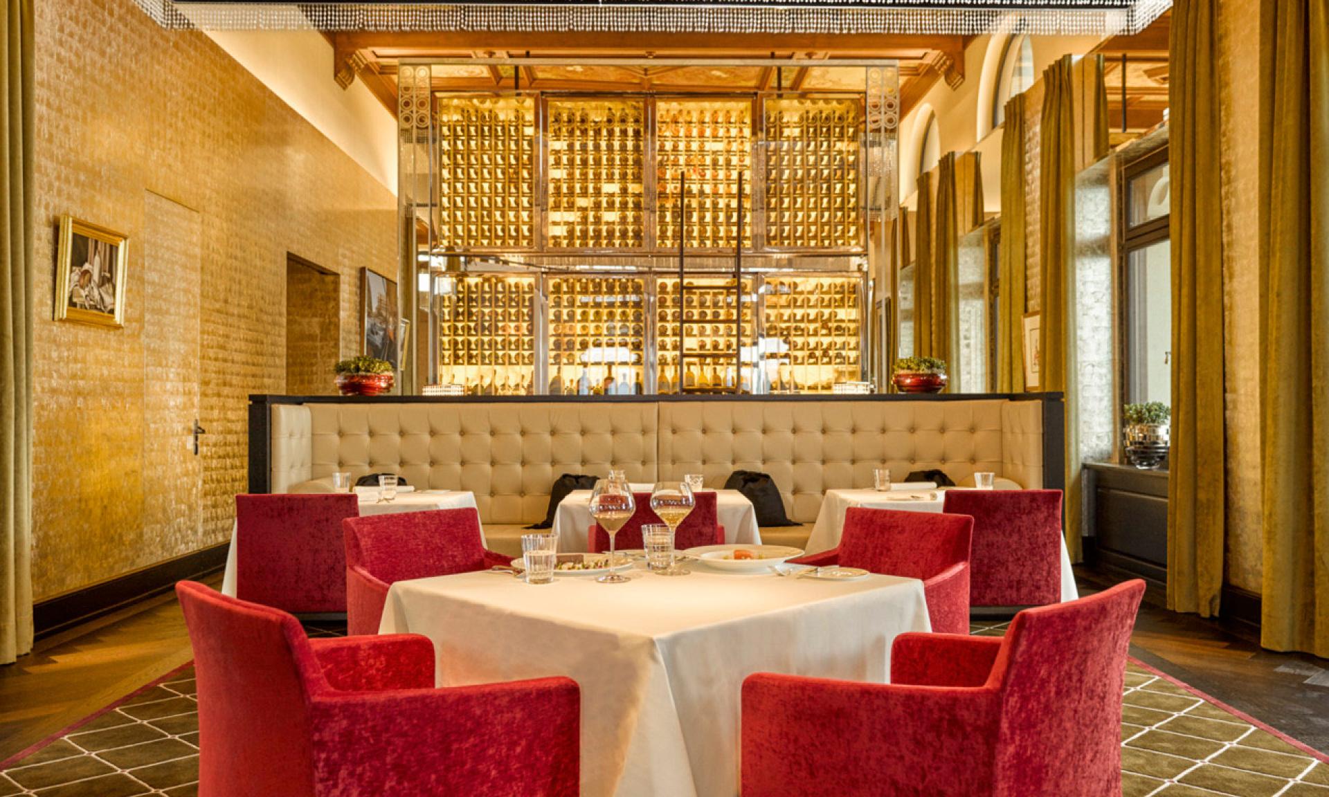 Elegante ristorante a Lugano con tavoli rotondi drappeggiati di stoffe bianche, circondati da sedie rosse. Un'esposizione di vini in vetro e toni dorati decorano lo spazio, con grandi finestre e soffitti alti che creano un'atmosfera lussuosa.
