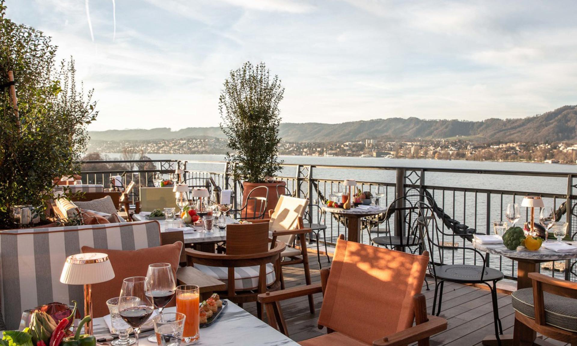 Terrazza ristorante all'aperto a Lugano con vista sul lago e sulle colline lontane, con tavoli apparecchiati, sedie imbottite e varie bevande e pasti alla calda luce naturale.