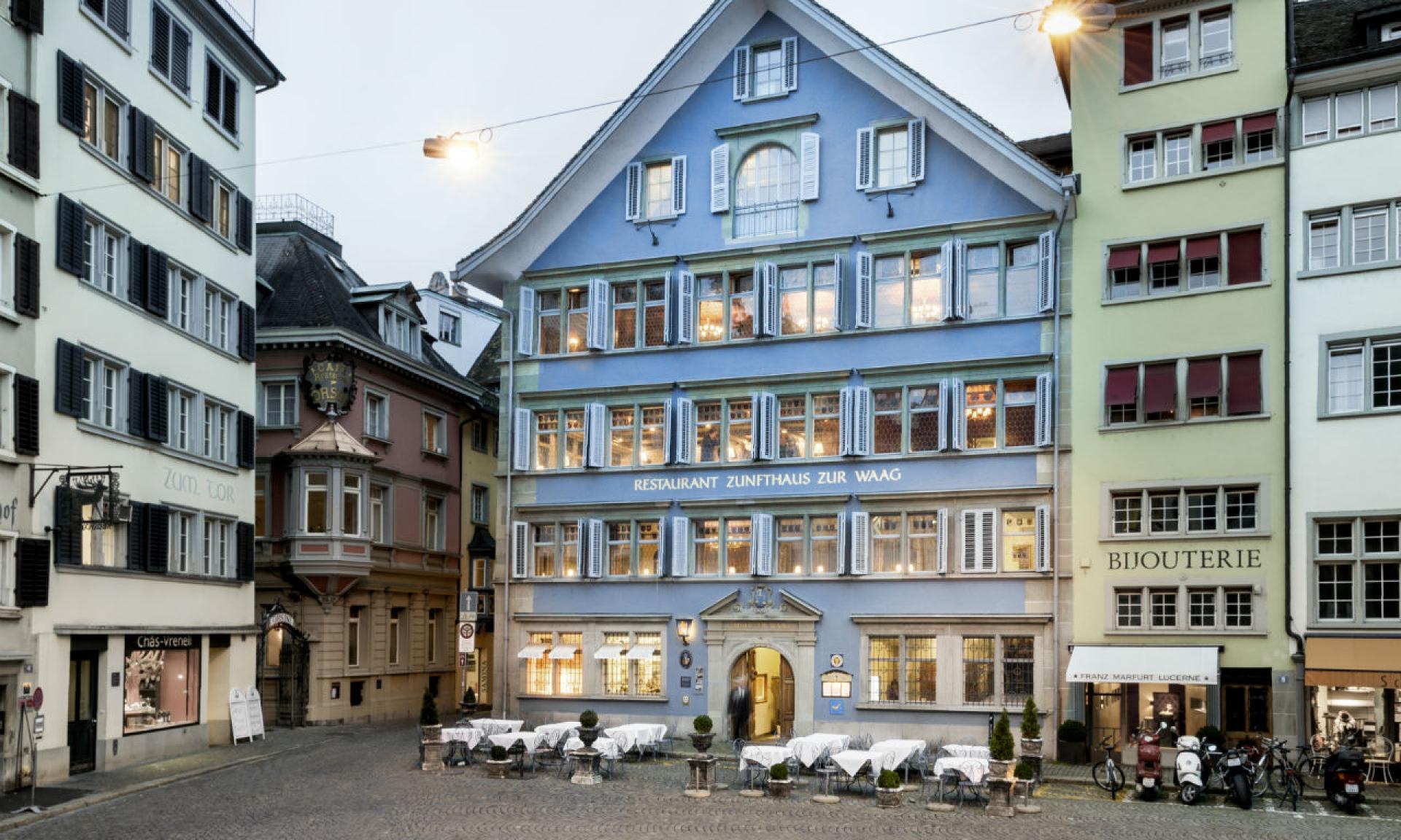 A blue three-story building with white trim houses Restaurant Zunfthaus zur Waag. Outdoor tables are set on a cobblestone plaza, surrounded by other colorful, old-style buildings in a European city.