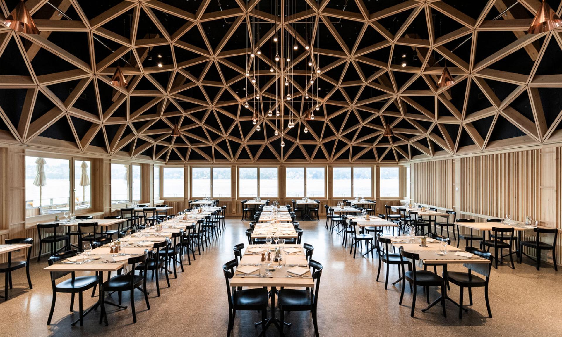 Un'ampia sala da pranzo del ristorante di Lugano con file di tavoli e sedie, allestita per gli ospiti. Il soffitto è caratterizzato da un intricato disegno geometrico in legno e le grandi finestre sul retro offrono una vista sull'acqua e sugli ombrelloni all'aperto.