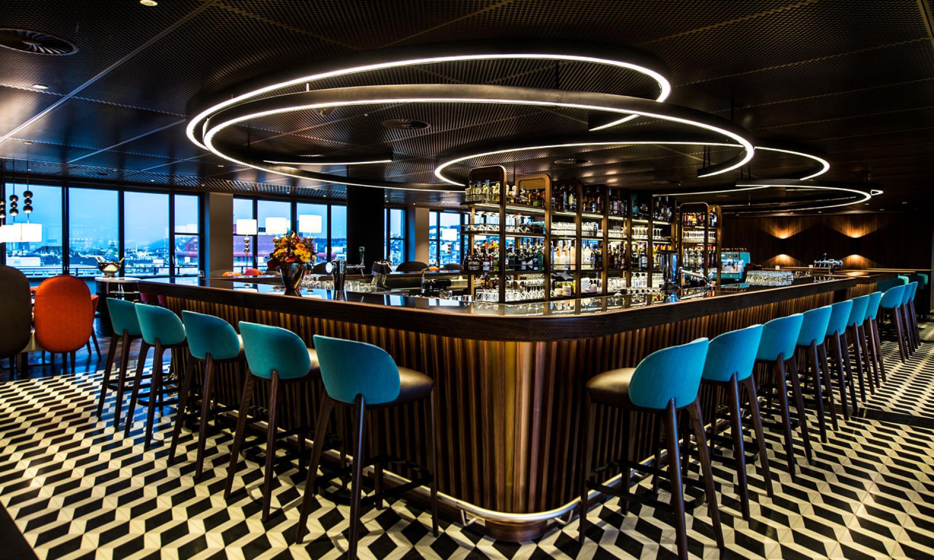 A modern bar with teal barstools, geometric black-and-white patterned floor, circular LED ceiling lights, and shelves of liquor bottles, set in a dimly lit space with large windows and city views.