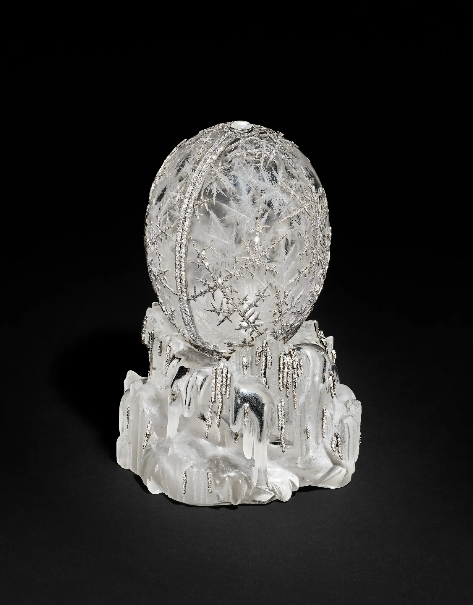 Una sfera di cristallo trasparente con intricati motivi incisi in stile Fabergé poggia su una base che ricorda il ghiaccio lucido che scorre o l'argento che si scioglie, il tutto su uno sfondo nero.
