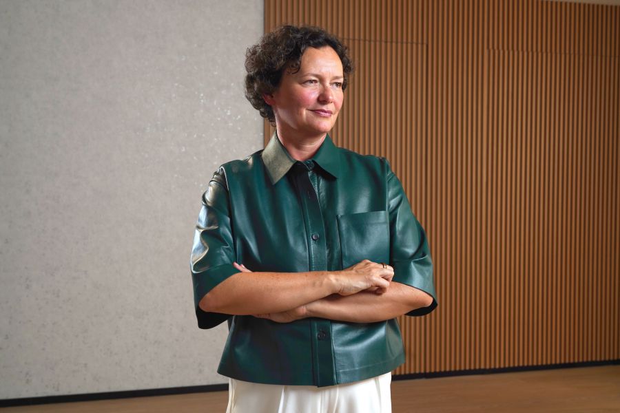 Una persona con i capelli corti e ricci, che indossa una camicia di pelle verde scuro a maniche corte e pantaloni bianchi, sta in piedi con le braccia incrociate in una stanza moderna con pannelli di legno e una parete dalla texture chiara, che incarna lo stile del Max Mara Art Prize.