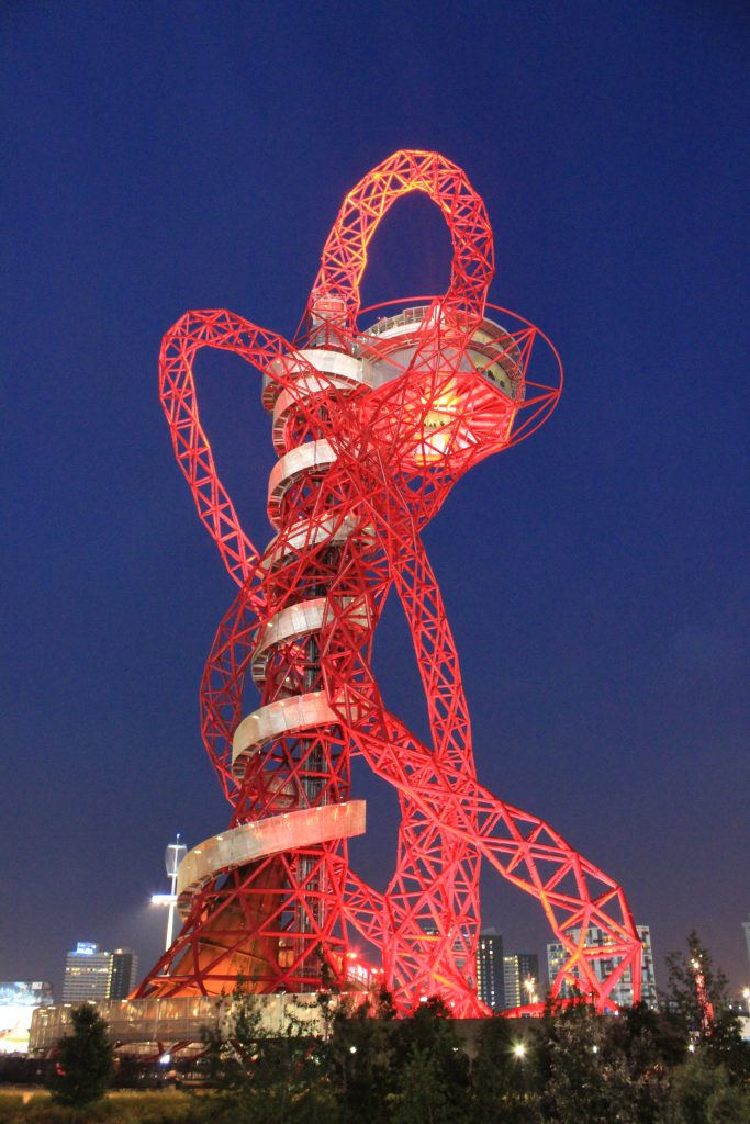 L'ArcelorMittal Orbit, un'opera dei giochi olimpici, è una torre di osservazione e uno scivolo tortuosi in acciaio rosso nel Parco Olimpico di Londra, illuminati di notte contro un cielo blu intenso con gli edifici della città sullo sfondo.