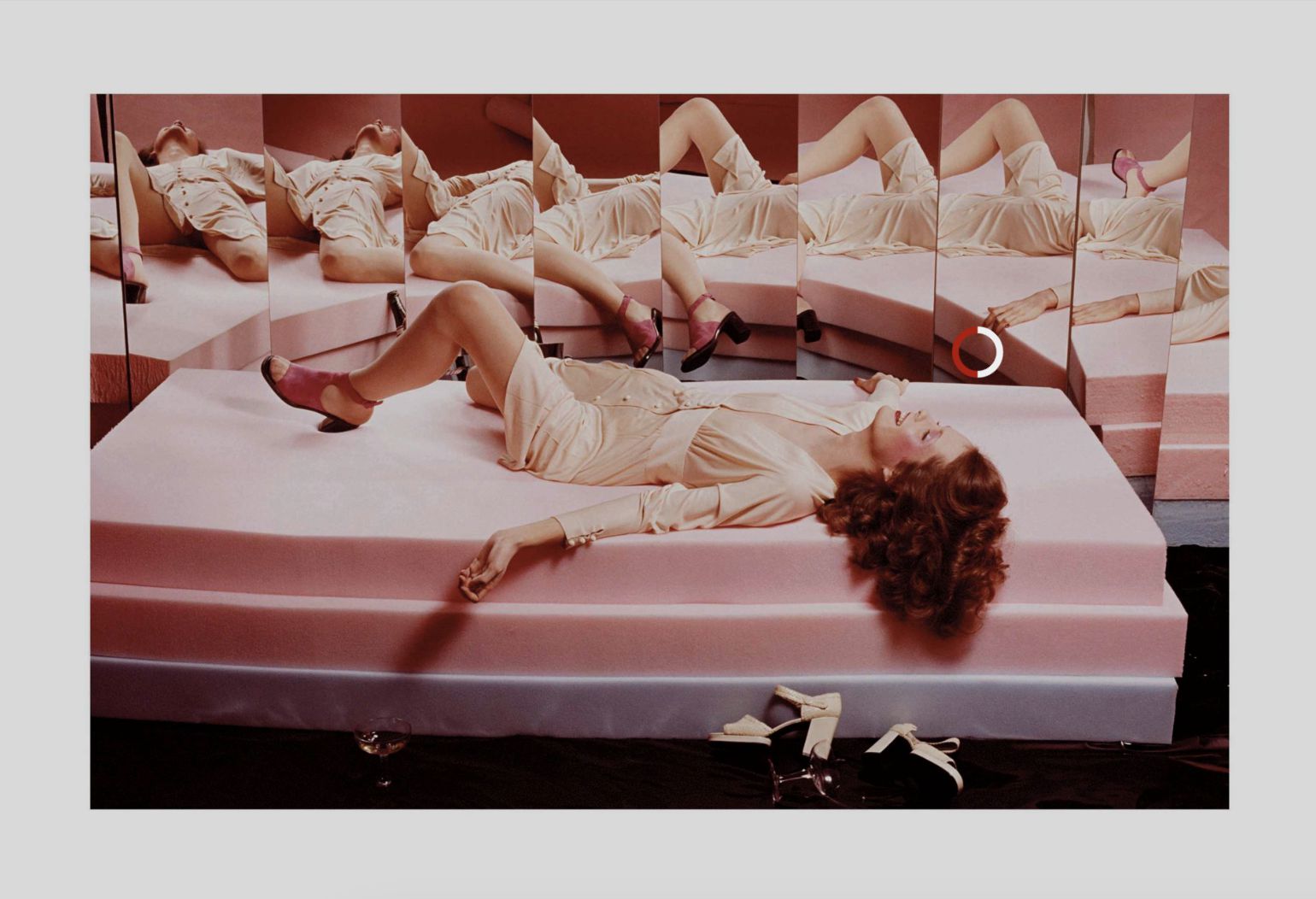 Una donna in abito beige giace su piattaforme di gommapiuma rosa, circondata da specchi che riflettono la sua immagine come un'installazione di Armani Arte. Le sue gambe sono incrociate, un braccio è sollevato e i tacchi alti rosa spiccano; scarpe bianche sono appoggiate sul pavimento vicino.