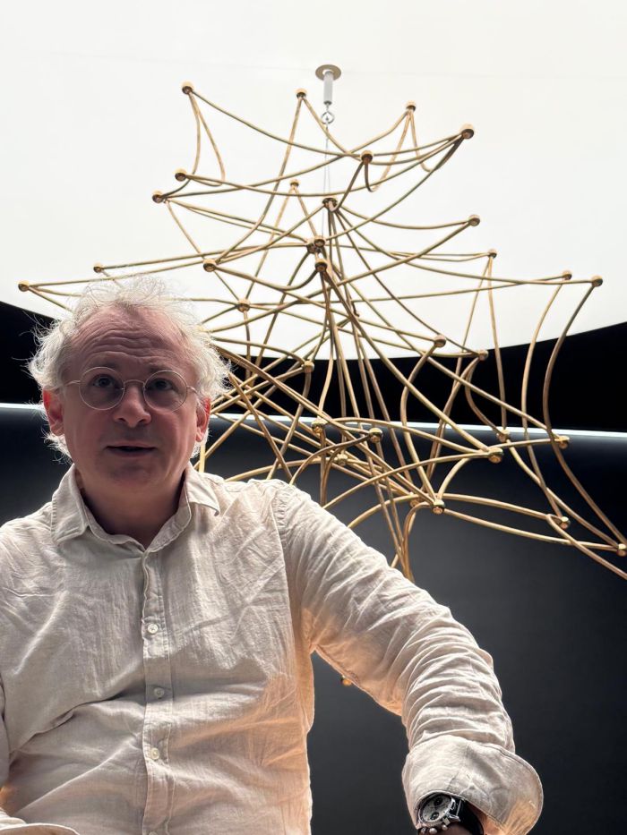 Un uomo con gli occhiali e i capelli bianchi, che indossa una camicia chiara, siede di fronte a una scultura geometrica, simile a una ragnatela, fatta di aste d'oro, su uno sfondo bianco e nero a contrasto.