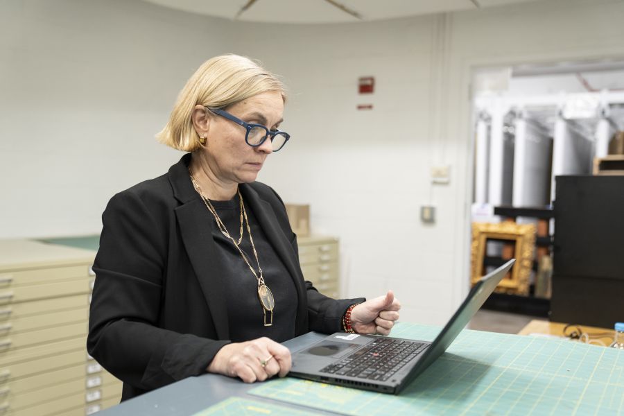 Una donna con i capelli biondi e gli occhiali, vestita di nero, è in piedi su un tavolo da lavoro che utilizza un computer portatile in un ufficio o in uno studio, circondata da cassetti e oggetti incorniciati - la sua attenzione ricorda un moderno Michelangelo scoperto al lavoro.