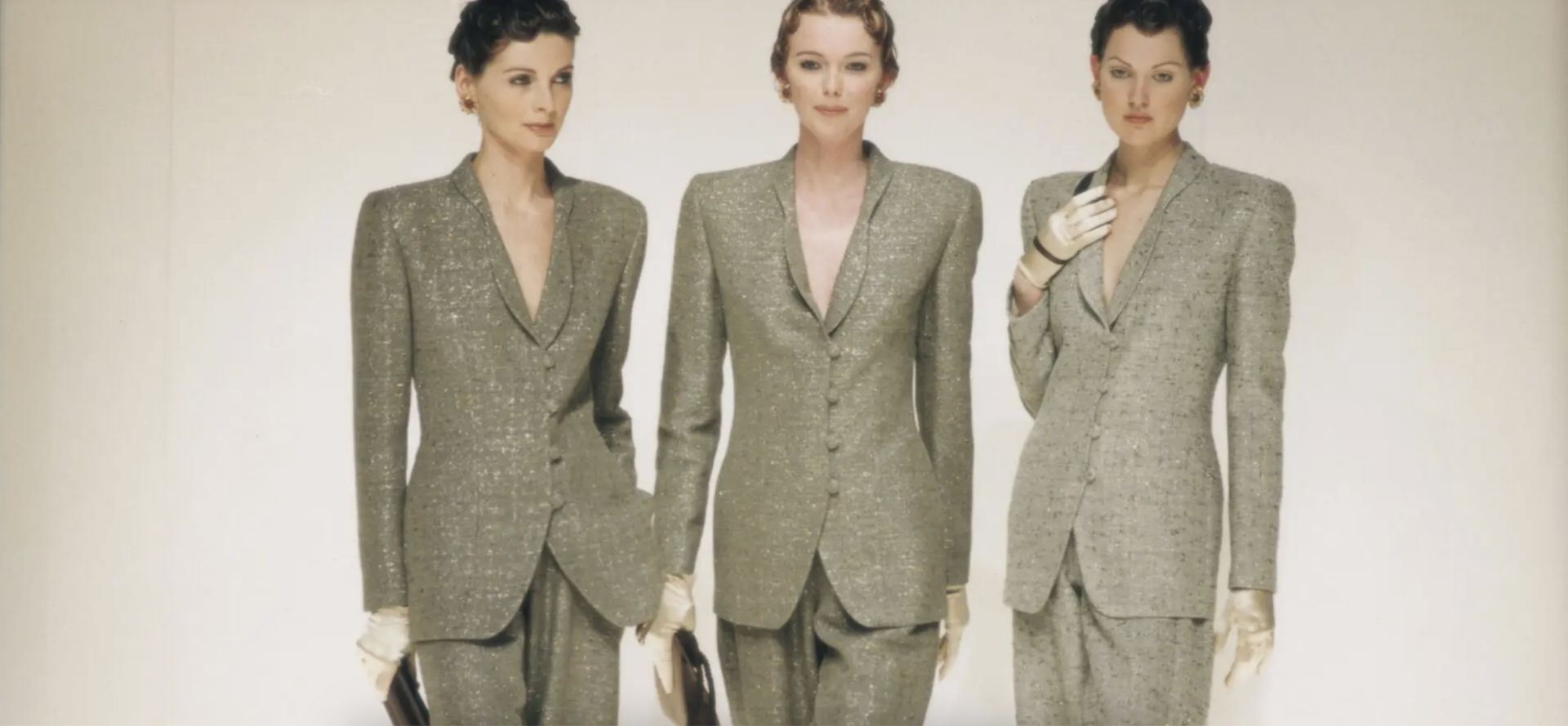 Tre donne affiancate in tailleur Armani arte grigio chiaro con spalle strutturate, guanti bianchi e pochette scure, su uno sfondo semplice. I loro capelli sono corti ed eleganti.