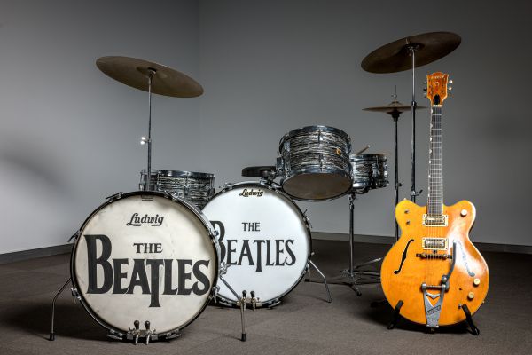 Una batteria vintage con il logo "The Beatles" sulla grancassa e una chitarra elettrica gialla esposta su un supporto, il tutto sullo sfondo di una parete grigia e di un pavimento in moquette: un vero gioiello per ogni collezione rock.