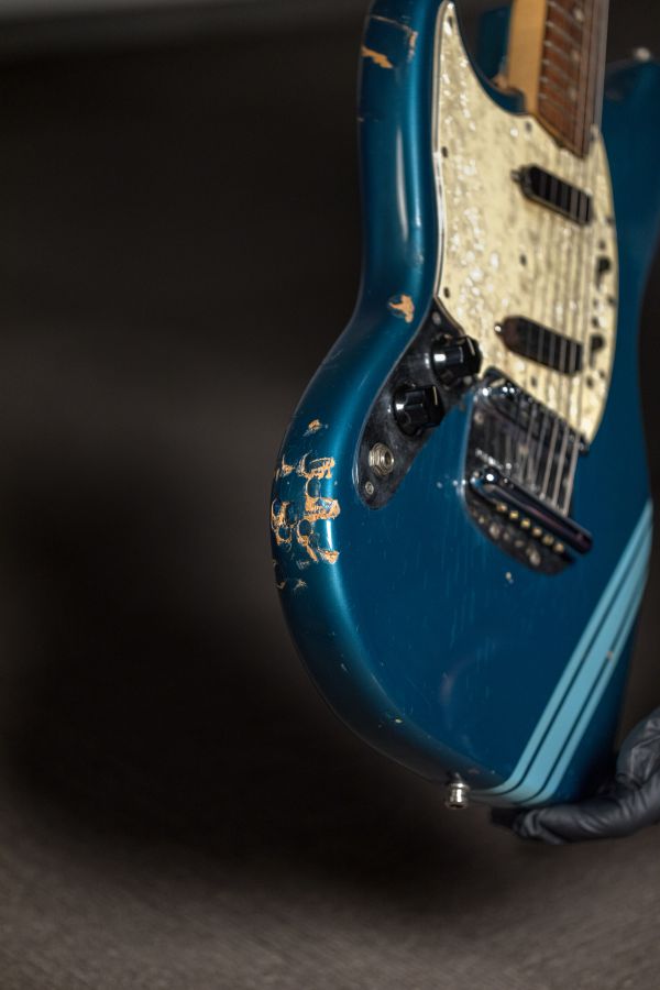 Primo piano di una chitarra elettrica blu usurata di una collezione rock, che mostra vernice scheggiata e graffi, con una mano guantata che tiene lo strumento su uno sfondo scuro.