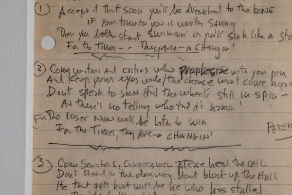 Un primo piano di una pagina scritta a mano su carta foderata, che mostra testi di canzoni numerati con parole cancellate e modificate: uno scorcio crudo di una collezione rock, dove la scrittura è leggermente disordinata e solo alcune parti del testo sono chiaramente leggibili.