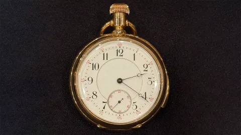 Un orologio da tasca d'oro della Collezione Beni 2025, con quadrante bianco, numeri neri e due lancette nere, si trova su uno sfondo di tessuto scuro. Un quadrante dei piccoli secondi è posizionato vicino alle ore 6.
