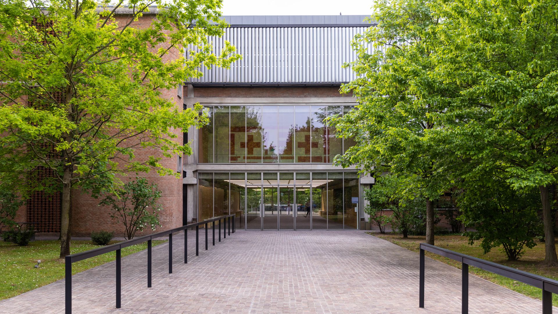 Un'ampia passerella fiancheggiata da ringhiere nere conduce a un moderno edificio in mattoni con ampie vetrate, uno spazio invitante che ricorda le sedi del Max Mara Art Prize, circondato da alberi verdi e prati rigogliosi su entrambi i lati.