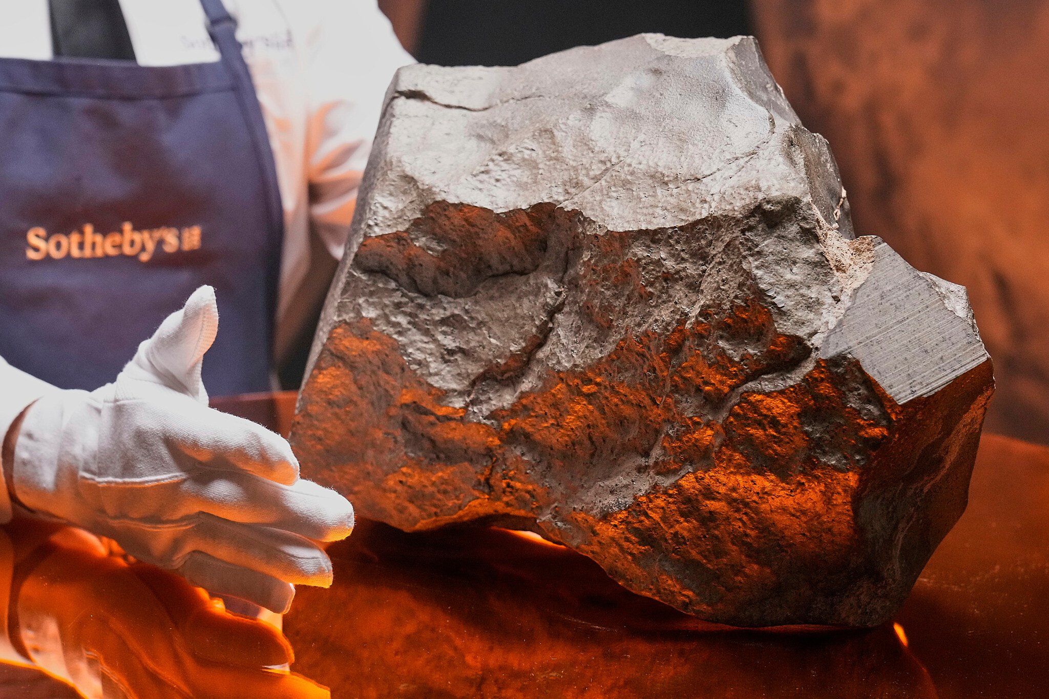Una persona che indossa guanti bianchi e un grembiule da Sotheby's presenta una roccia grigia di grandi dimensioni e di forma irregolare - forse un meteorite aste record - esposta su una superficie riflettente con una calda illuminazione arancione.