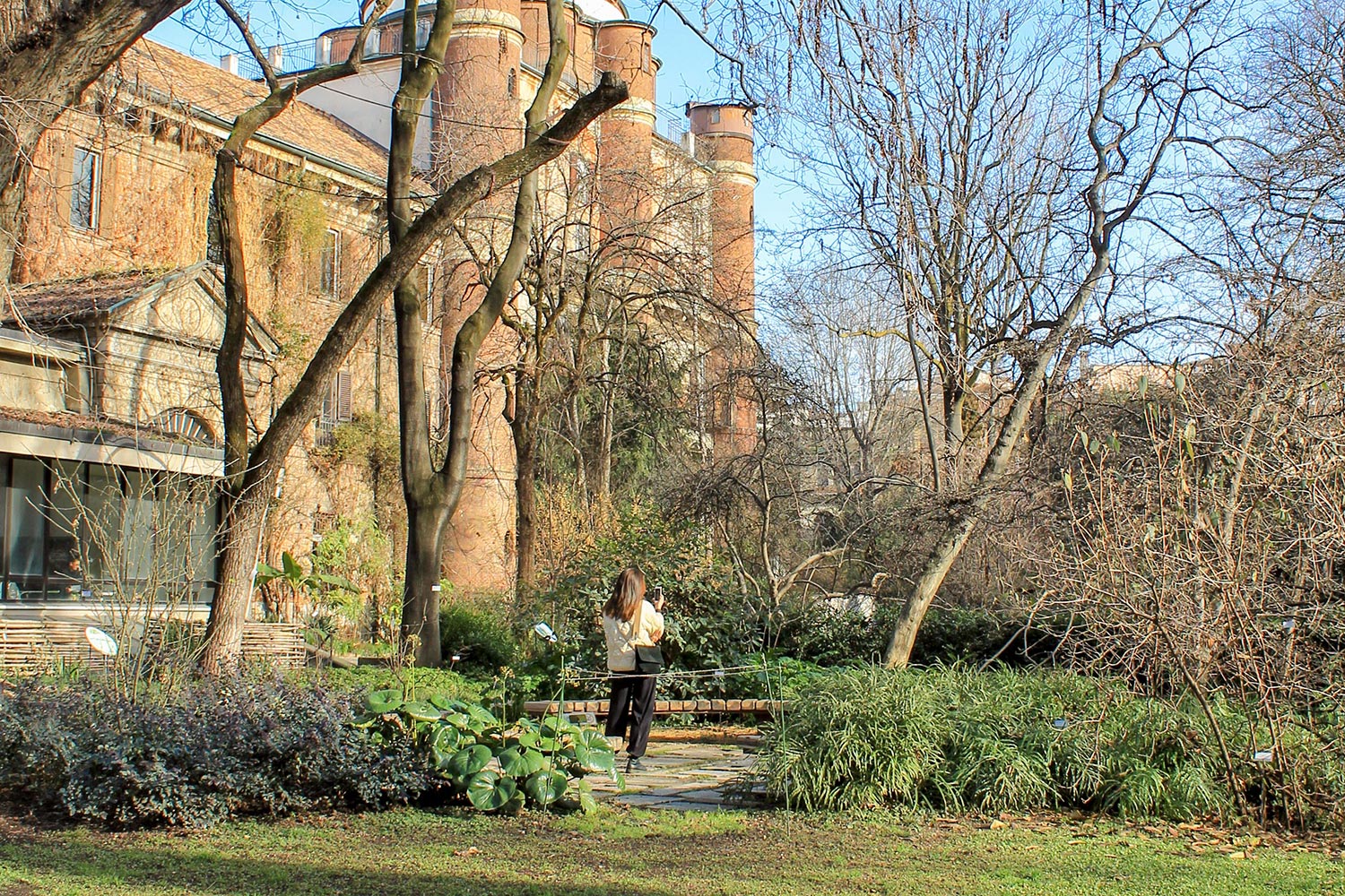 Una persona con i capelli lunghi cammina lungo un sentiero del giardino circondato da alberi e verde a Lugano, con un grande edificio in mattoni e diversi camini cilindrici sullo sfondo sotto un cielo azzurro e limpido.