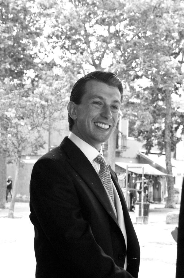 Un uomo in giacca e cravatta sorride calorosamente mentre stringe la mano all'aperto, con alberi ed edifici sfocati sullo sfondo. L'immagine è in bianco e nero.