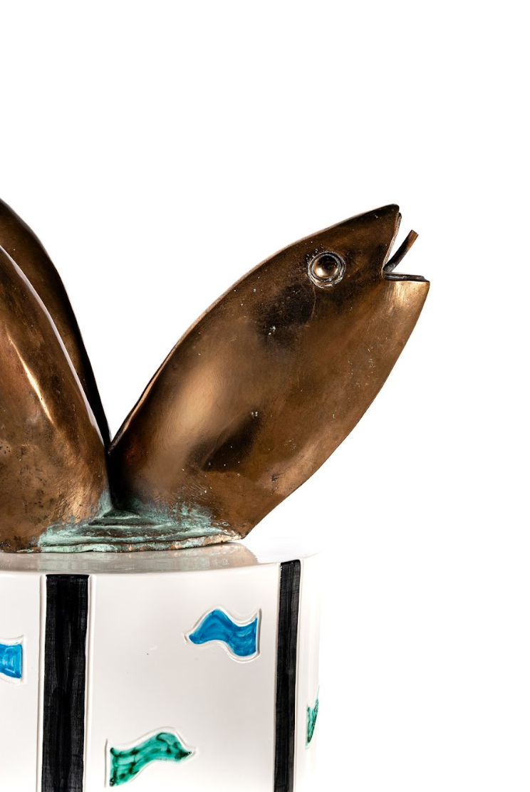Primo piano di una scultura in bronzo di un pesce con la bocca aperta, montata su una base bianca decorata con linee verticali nere e motivi ondulatori blu e verdi: un esempio straordinario di arte kruso. Lo sfondo è bianco.