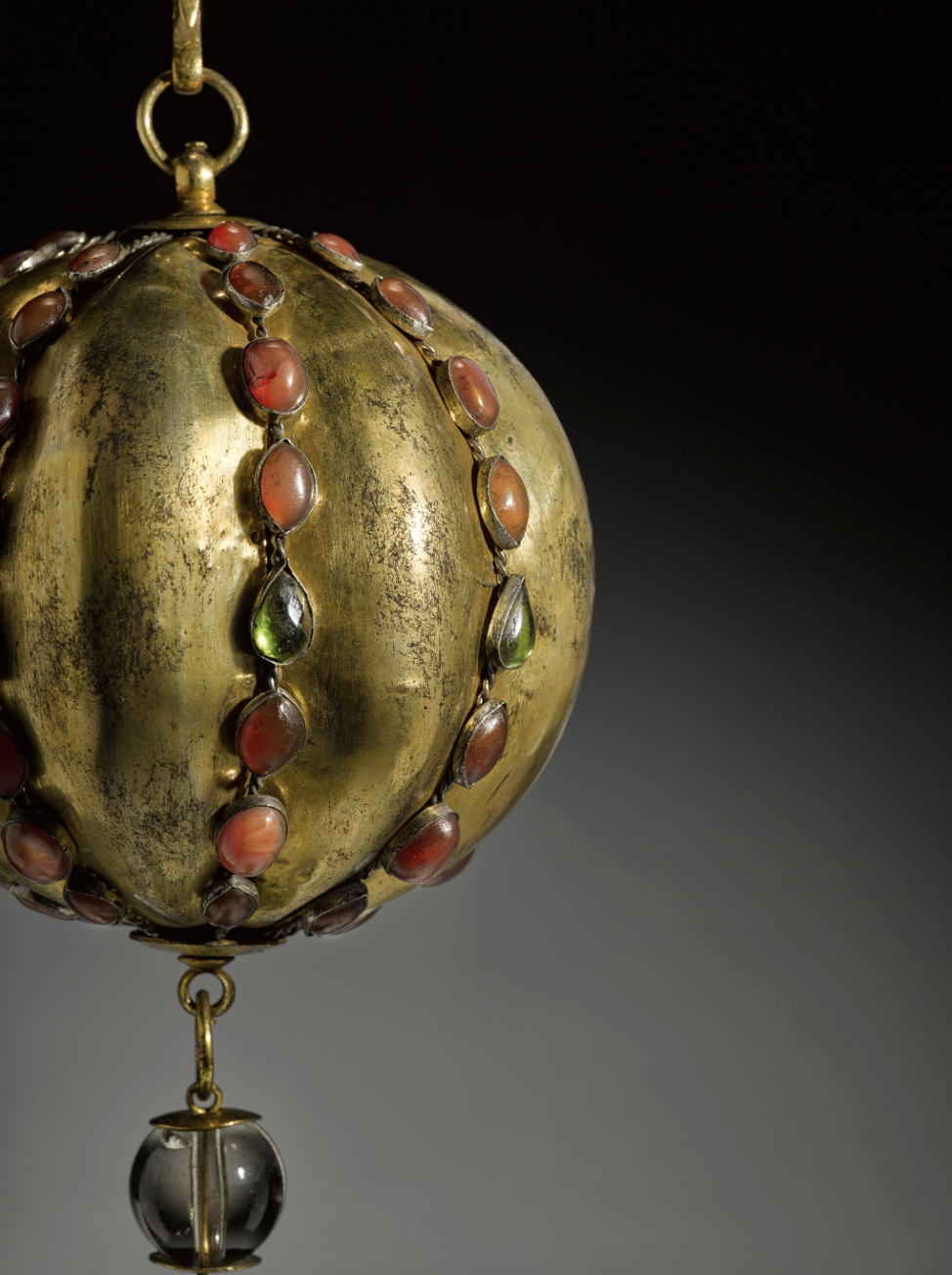 Ornamento sferico in oro antico di Arte Islamica Collezioni, decorato con gemme ovali rosse e verdi e un piccolo pendente in vetro che pende dalla parte inferiore, su uno sfondo scuro e sfumato.