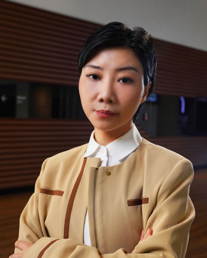 Una donna con i capelli corti e neri, che indossa una giacca beige su una camicia bianca, sta in casa con le braccia incrociate e guarda con sicurezza la macchina fotografica. Il pannello di legno sfocato fa pensare a un evento del Max Mara Art Prize.