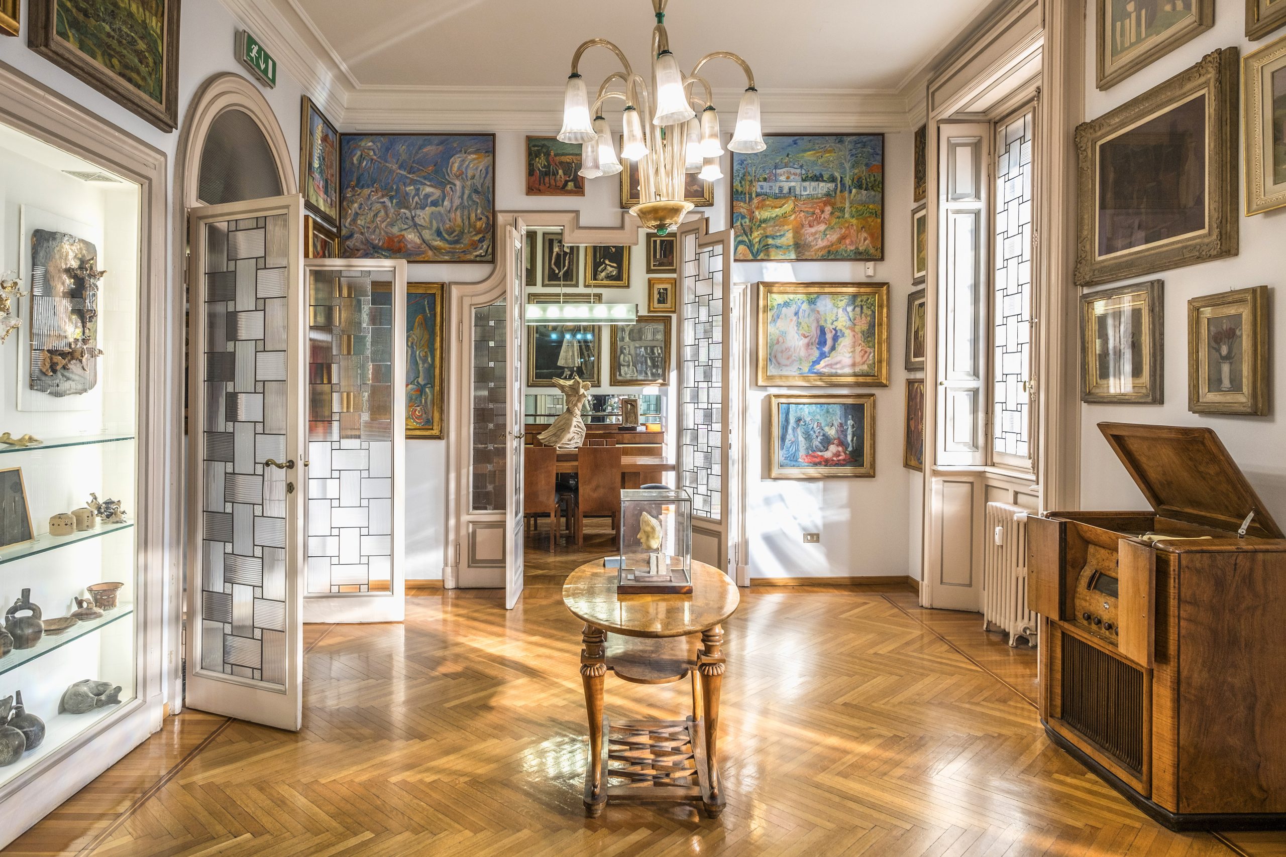 Una sala luminosa ed elegante a Lugano, con pavimenti in parquet, grandi finestre e pareti ornate di quadri. Al centro si trova un tavolo rotondo, con esposizioni d'arte, un lampadario e porte a vetri che conducono a un'altra stanza.
