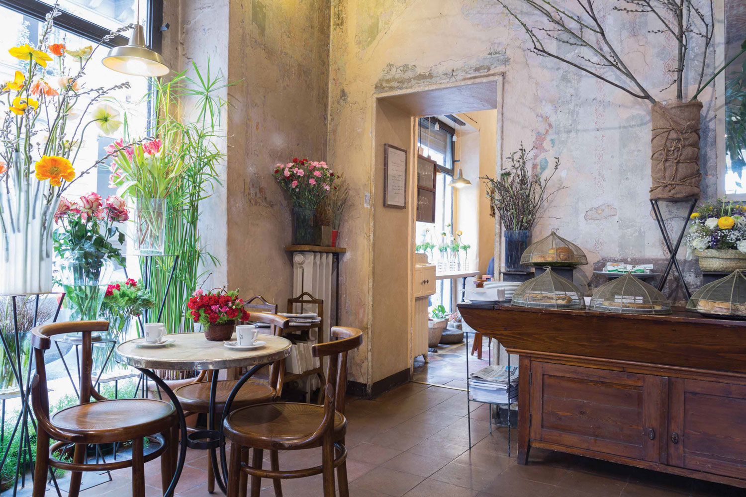 Un accogliente caffè di Lugano con sedie in legno e tavoli rotondi in marmo, circondati da alti vasi di fiori colorati. Le pareti rustiche e il bancone in legno espongono i dolci sotto cupole di vetro, mentre la luce del sole filtra dalle grandi finestre.