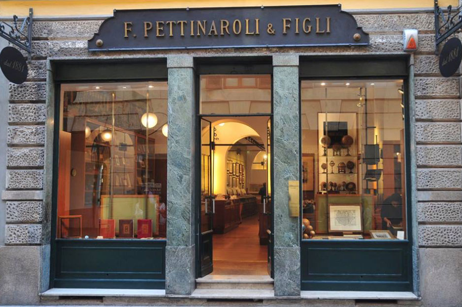 Veduta stradale della vetrina "F. Pettinaroli & Figli", caratterizzata da ampie vetrine con articoli di cartoleria e antiquariato, che ricordano i classici negozi di Lugano, con una porta a vetri al centro e un'insegna in legno sopra l'ingresso.