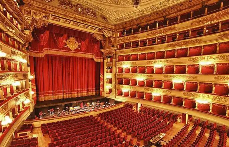 Ornato teatro dell'opera di Lugano con poltrone di velluto rosso, balconi multipli e un grande palcoscenico incorniciato da un elaborato sipario rosso e decorazioni dorate; gli orchestrali si stanno sistemando nella buca dell'orchestra.