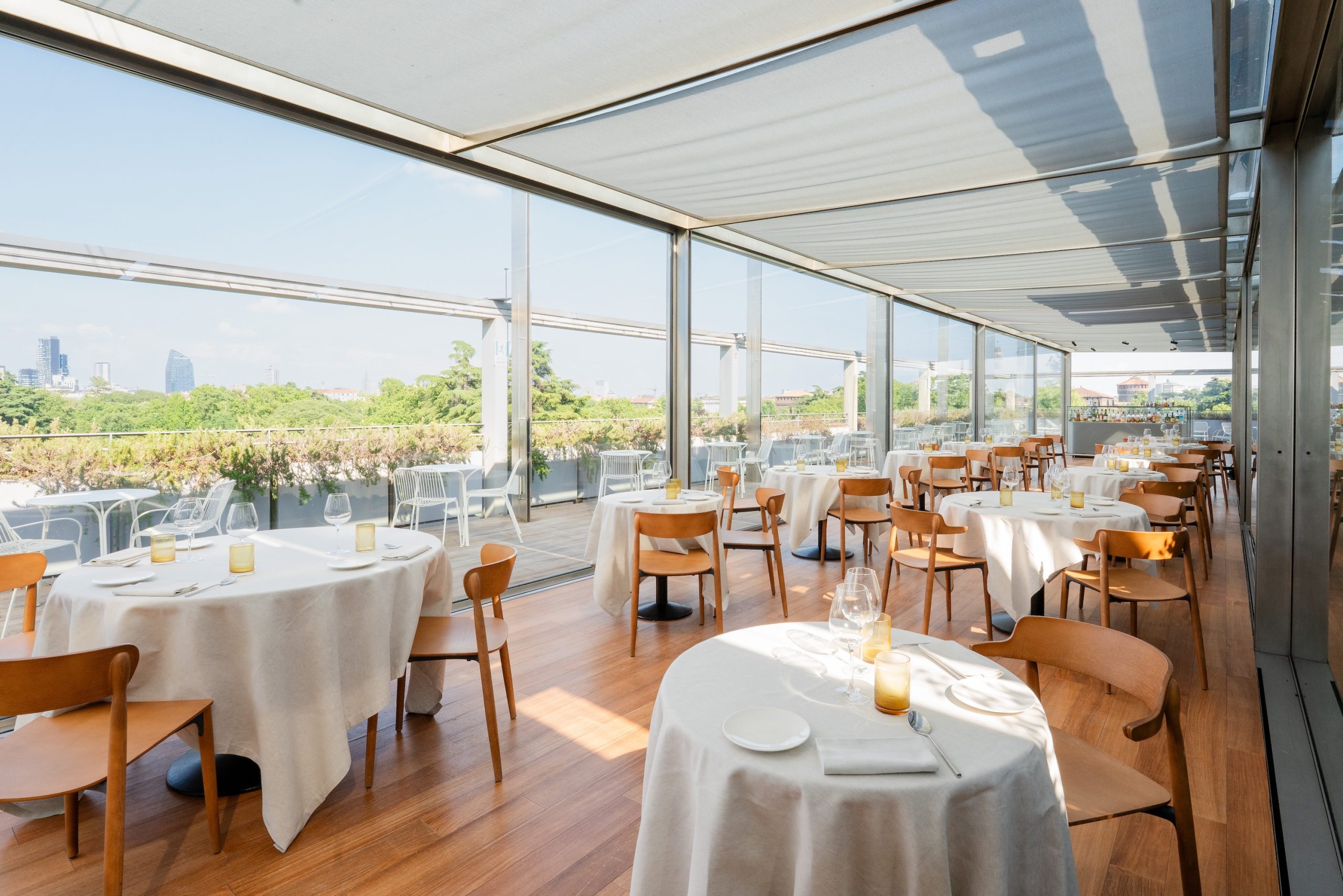 Un ristorante luminoso e moderno a Lugano con pavimenti in legno, tavoli rotondi coperti da tovaglie bianche e sedie in legno. Grandi finestre si aprono su una terrazza con vista sul verde e sulla città sotto un cielo azzurro.