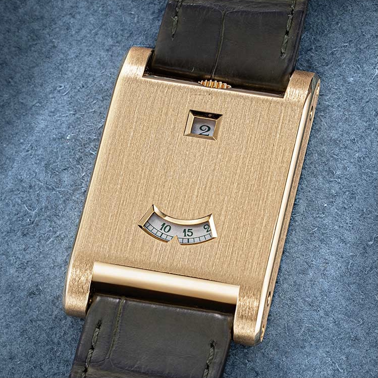 Orologio da polso d'oro con cassa rettangolare spazzolata, cinturino in pelle nera, piccola finestrella quadrata con la cifra "2" vicino alla parte superiore e finestrella curva con i minuti su sfondo blu, incastonata su una superficie morbida blu.