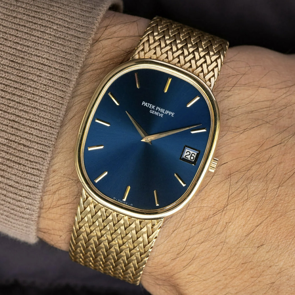 Un orologio da polso Patek Philippe in oro con quadrante blu, indici in oro e datario a ore 3 è indossato al polso di una persona con un maglione beige. L'orologio ha un bracciale d'oro intrecciato.