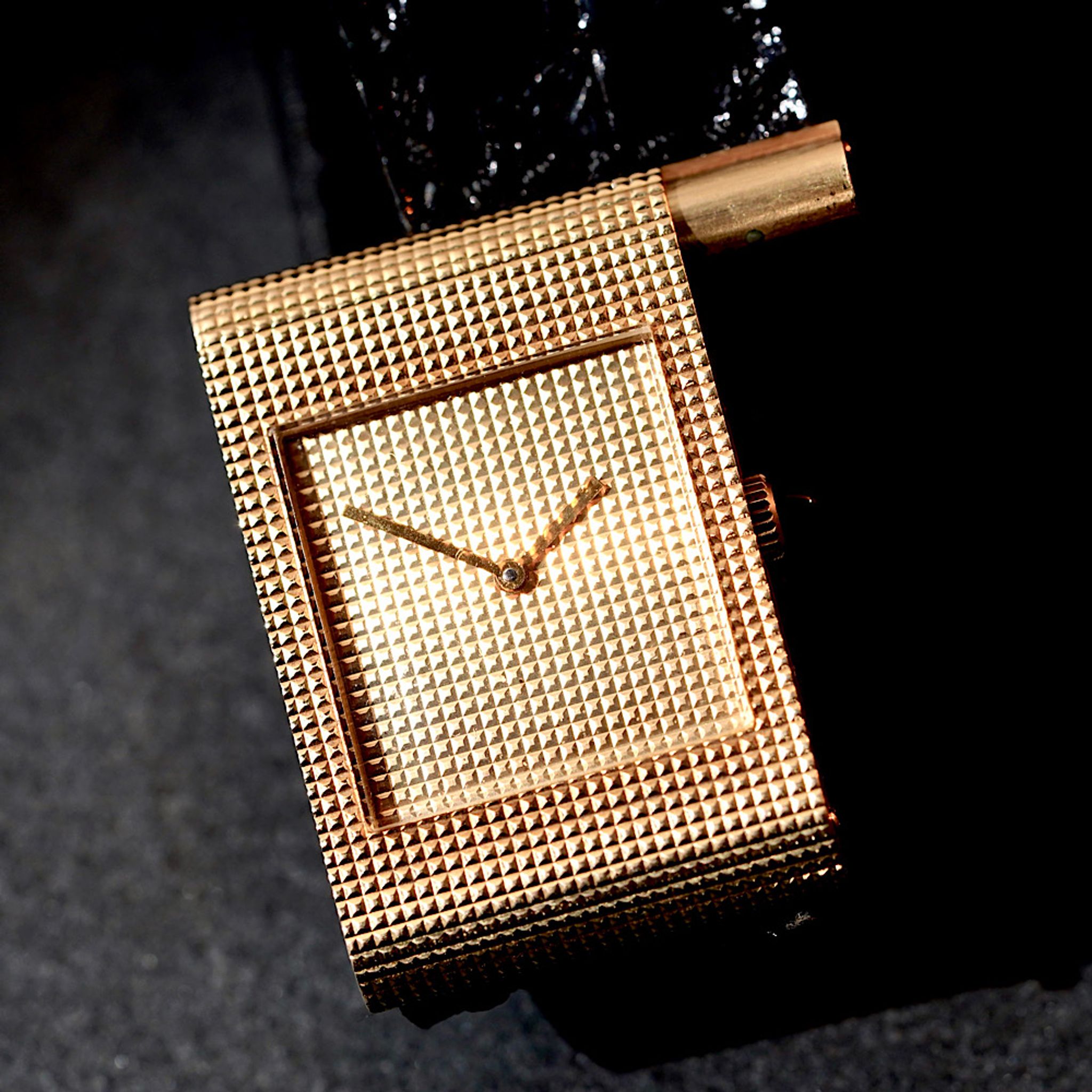 Orologio da polso d'oro con quadrante quadrato testurizzato e motivo a griglia e lancette d'oro abbinate, su un cinturino nero su sfondo scuro.