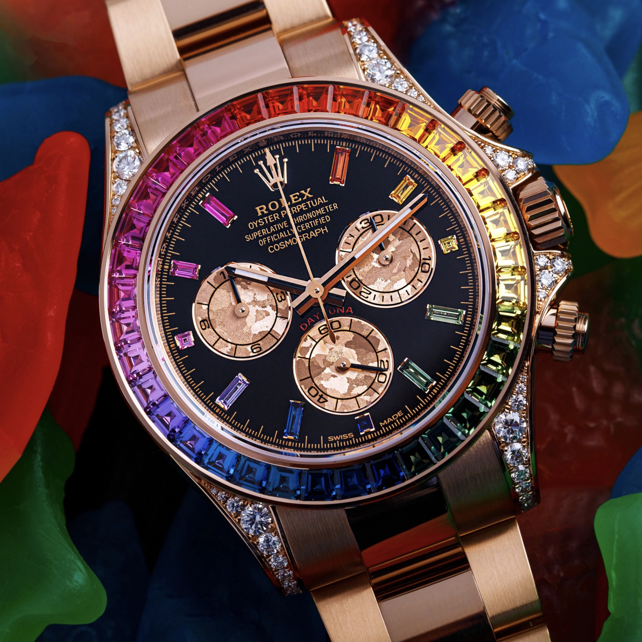Orologio Rolex d'oro con quadrante nero e tre contatori, caratterizzato da una lunetta con gemme multicolori disposte in un motivo arcobaleno, circondata da orsetti gommosi colorati.