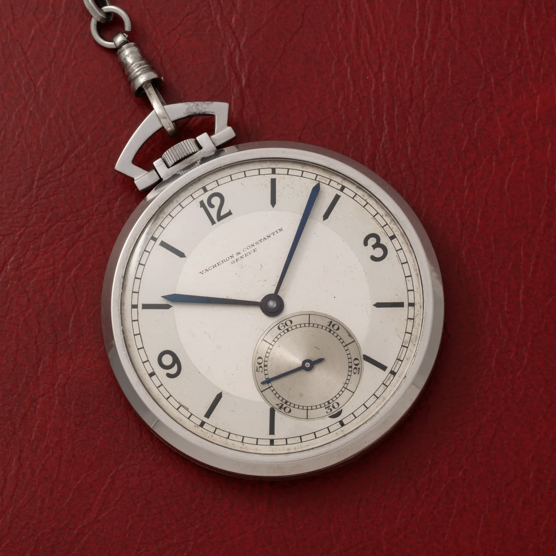 Un orologio da tasca d'argento con lancette blu e quadrante dei piccoli secondi poggia su una superficie rossa testurizzata. Il quadrante dell'orologio indica l'ora 12:14 e reca la dicitura "Vacheron & Constantin".