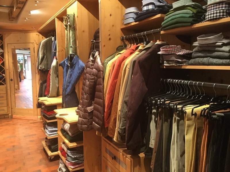 Un negozio di abbigliamento ben organizzato, con scaffali e rastrelliere in legno che espongono camicie, giacche, maglioni e pantaloni piegati, perfetti per un weekend milanese. Il pavimento piastrellato conduce a una porta sul retro che si apre su un'altra stanza.