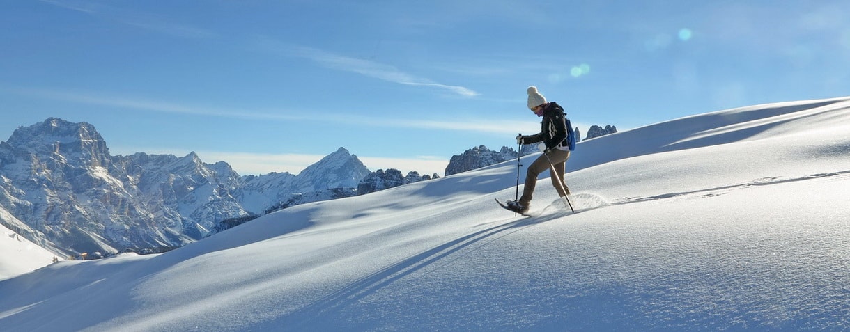 Una persona che indossa abiti invernali cammina in salita nella neve alta con le racchette da neve, circondata da montagne innevate sotto un cielo azzurro e limpido: una fuga ideale per il weekend a Milano.