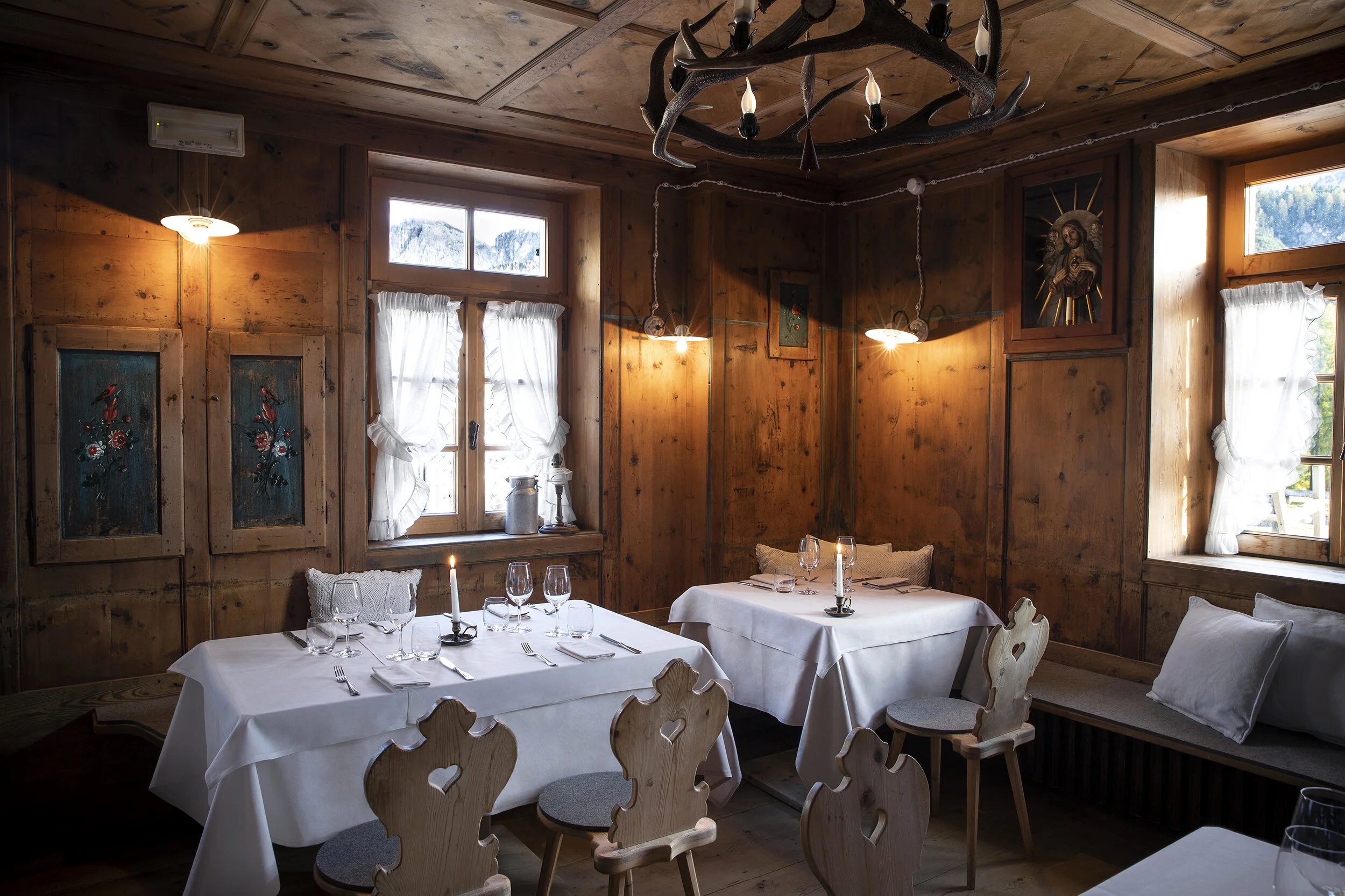 Un'accogliente sala da pranzo rustica con pareti rivestite in legno, sedie in legno intagliato e tavoli a lume di candela crea la scena per un perfetto weekend milanese. Una luce soffusa filtra dalle finestre, creando un'atmosfera calda e invitante sotto un lampadario di corna.