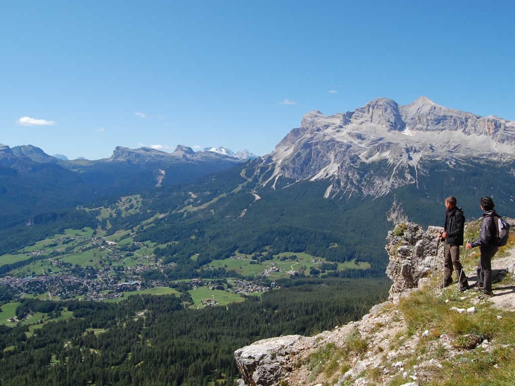 Due escursionisti si trovano su una sporgenza rocciosa che domina una valle verdeggiante e un'incantevole città - una fuga ideale per un weekend a Milano - circondata da colline boscose e cime montuose drammatiche e illuminate dal sole sotto un cielo azzurro e limpido.