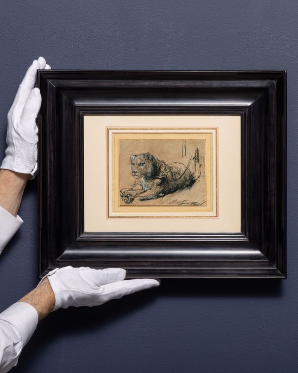 Una persona che indossa guanti bianchi tiene in mano uno schizzo incorniciato di un leone sdraiato, che ricorda gli studi di Rembrandt sul leone, contro una parete grigia semplice. L'opera è circondata da una stuoia bianca e da una cornice scura e larga.