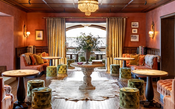 Un lussuoso salotto con sedie di velluto, tavoli rotondi e pouf in legno, perfetto per un weekend a Milano. Un tavolo rotondo con una composizione floreale si trova su un tappeto, mentre le tende e il lampadario aggiungono eleganza alla stanza caldamente illuminata.