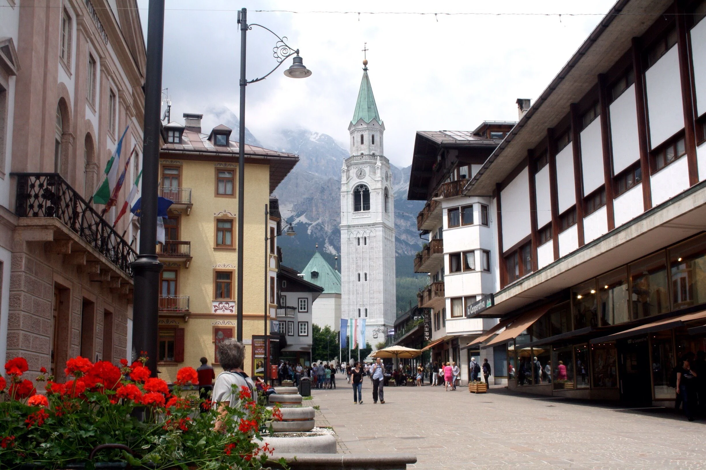 Una pittoresca piazza della città, perfetta per un weekend milanese, con fiori rossi in primo piano, persone che passeggiano, edifici colorati e un alto campanile bianco con una guglia verde incastonata tra montagne nuvolose.