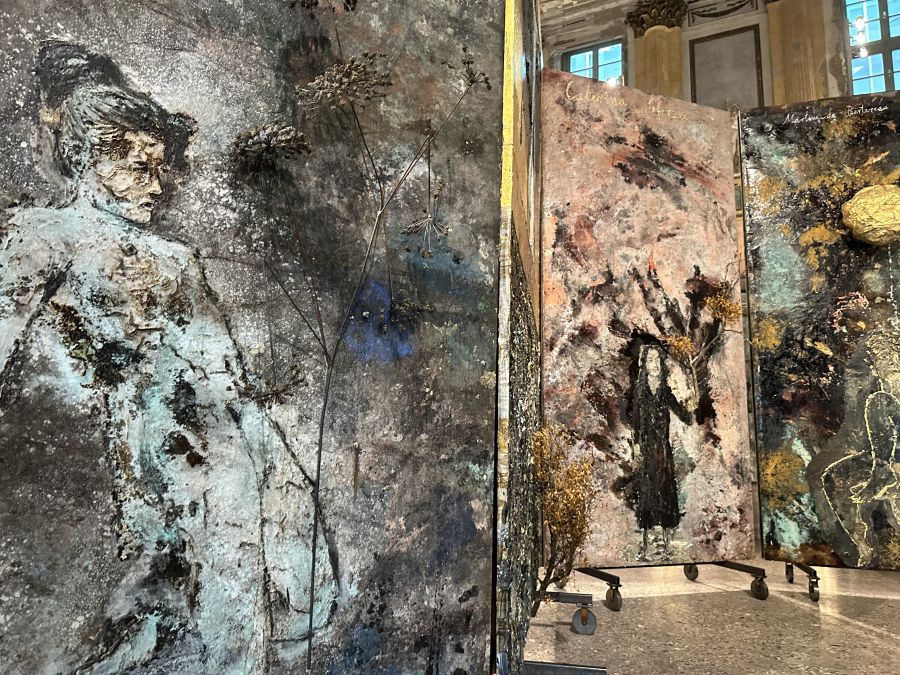 Pannelli a tecnica mista con disegni strutturati, astratti e figurativi che ricordano Anselm Kiefer sono esposti in uno spazio della galleria ben illuminato con alte finestre e un'architettura ornata sullo sfondo.
