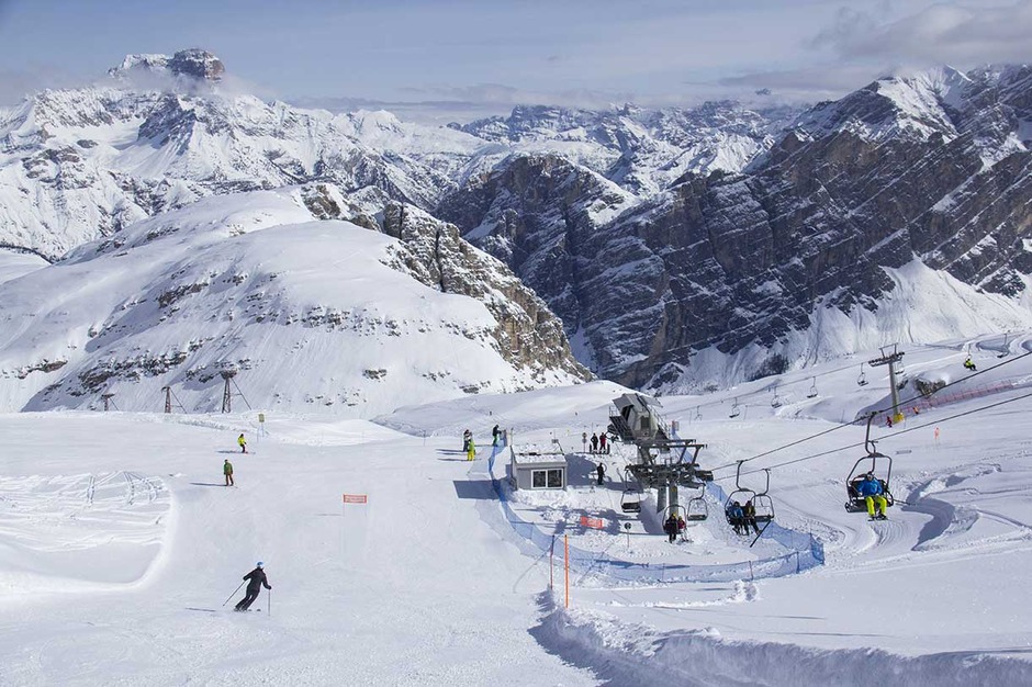 Sciatori e snowboarder si godono una pista di montagna innevata con seggiovie, circondata da cime alte e aspre sotto un cielo parzialmente nuvoloso: uno scenario vivace perfetto per un weekend di fuga a Milano.