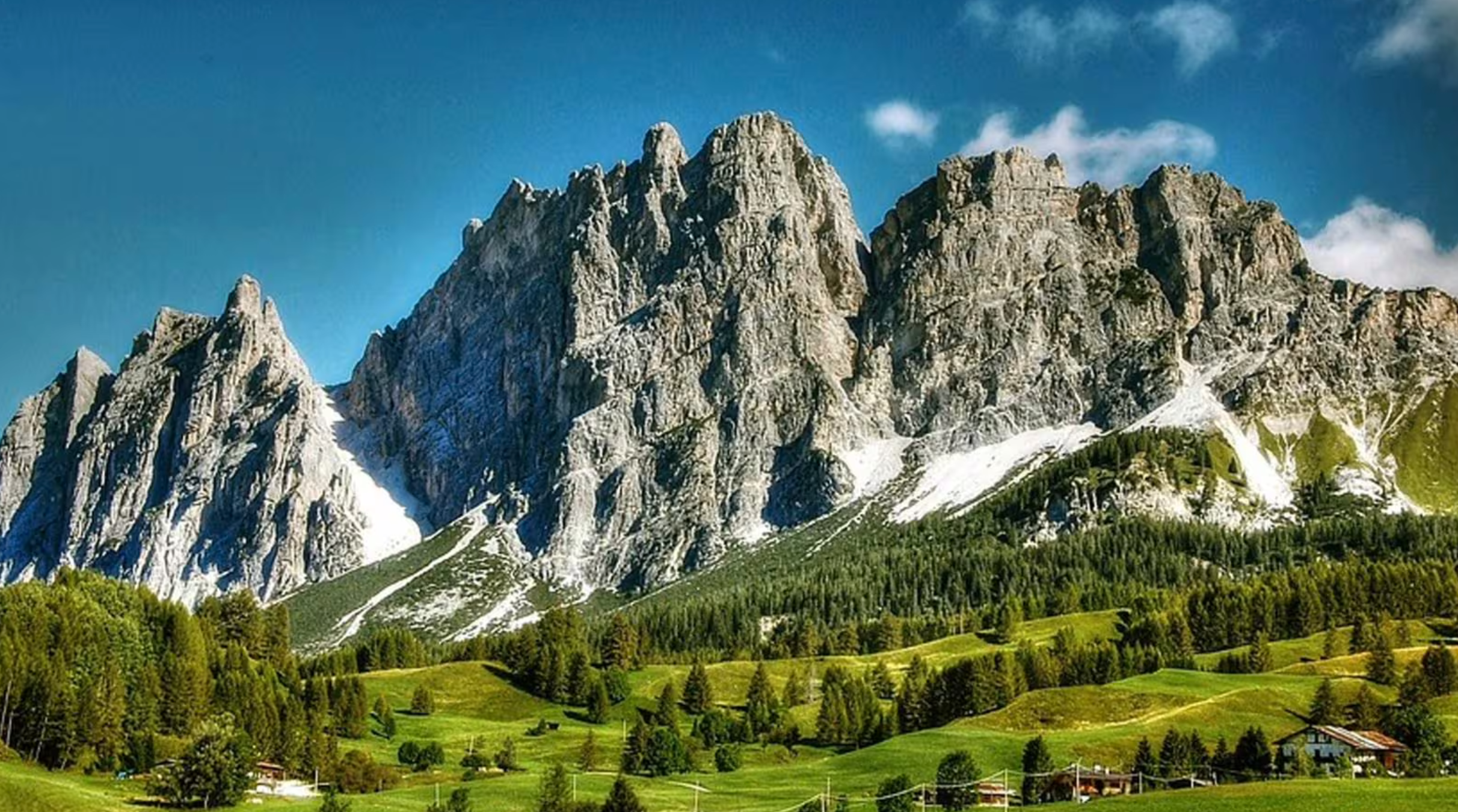 Montagne ripide e rocciose si ergono sopra colline verdi e lussureggianti, punteggiate di alberi e piccole case - una fuga ideale per un weekend rilassante a Milano - sotto un cielo azzurro e limpido con nuvole sparse e macchie di neve persistenti vicino ai pendii boscosi.