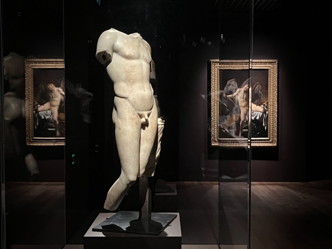 Una statua di marmo raffigurante un torso maschile nudo è esposta in una galleria poco illuminata, con due dipinti incorniciati di figure nude reclinate visibili sullo sfondo.