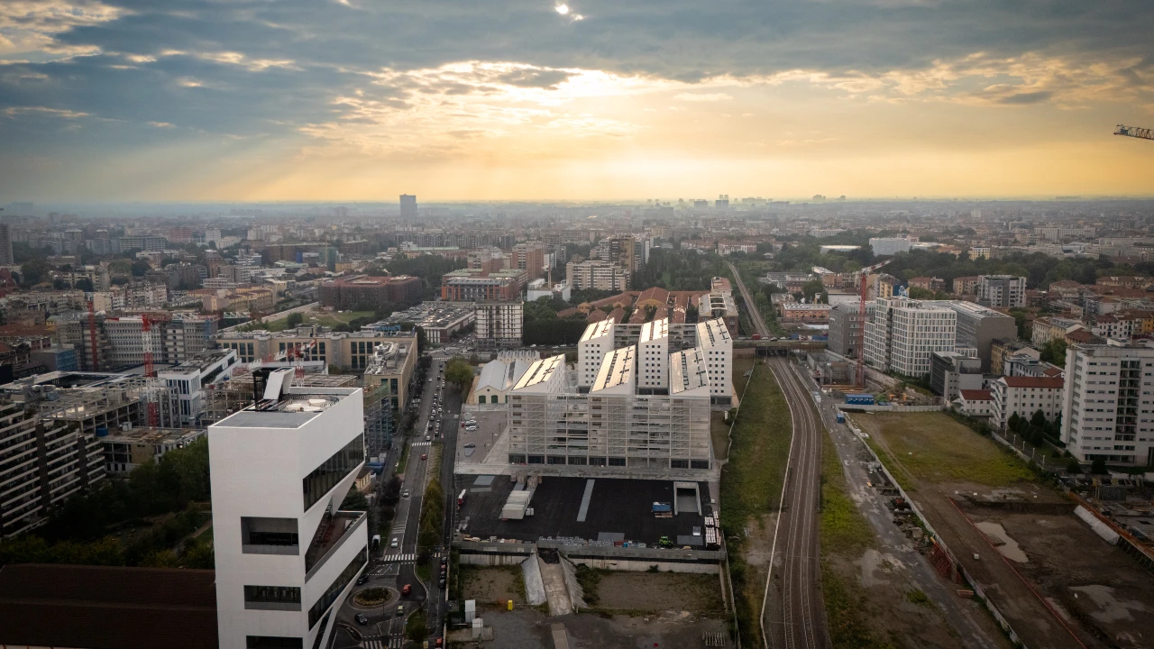 A cityscape at sunset with rays of sunlight piercing through clouds, illuminating modern buildings, roads, and train tracks, with a sprawling urban area extending toward the horizon.